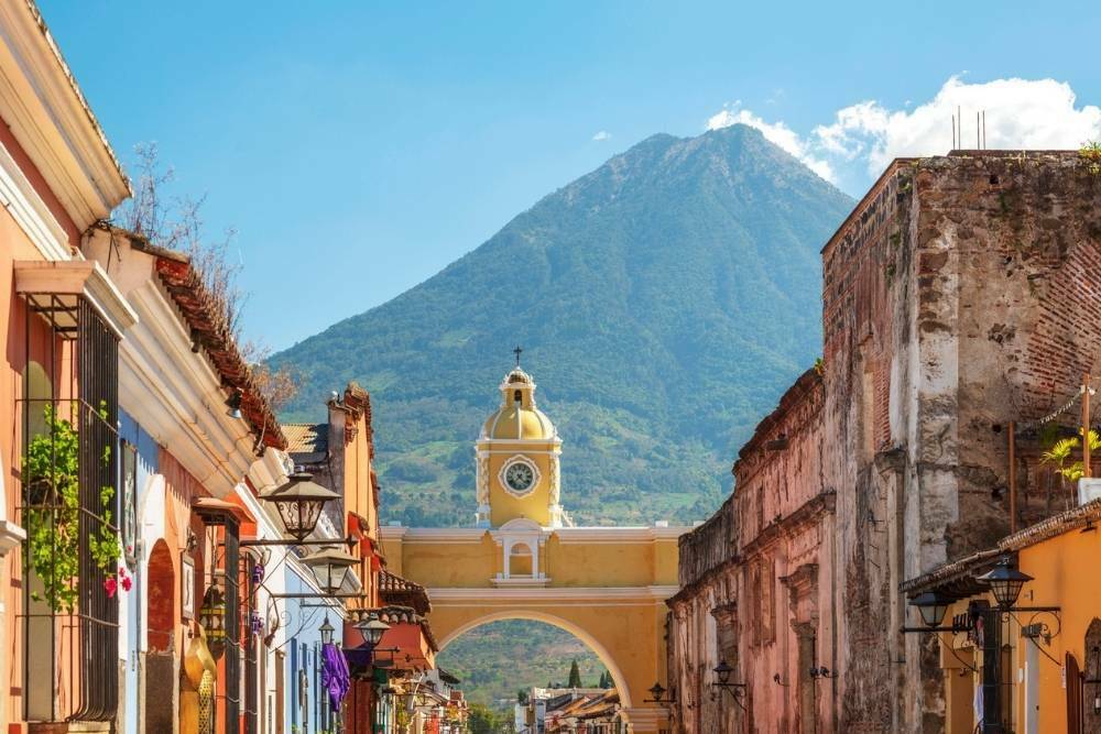 An image of one of the most beautiful places in Guatemala--Antigua Guatemala--with a close up view of a yellow arched bridge, the Santa Catalina Arch, with a volcano seen behind it, the Volcán de Agua