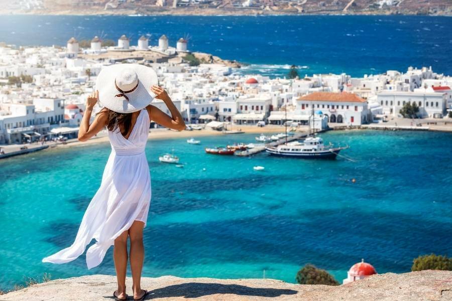 White Beach Dress Outfits for Greece - woman standing on cliff in Mykonos Greece wearing a white beach dress outfit with a white hat